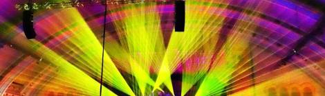 Distant shot of band on concert star with purple, green, and yellow lights at center