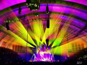 Distant shot of band on concert star with purple, green, and yellow lights at center